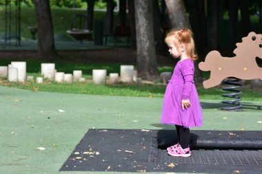 Profil görüntüsü, mor elbiseli, pembe sandaletli, çocuk parkında dikilen, ciddi bir ifadeyle ileriye bakan, parkta güneşli bir gün..