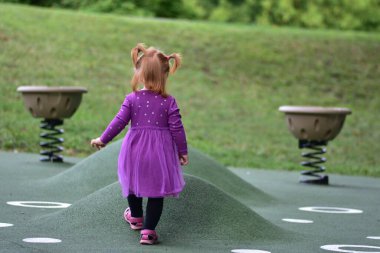 Kırmızı saçlı, mor elbiseli, pembe sandaletli bir kızın yeşil çocuk parkında yürüdüğü arka manzara oyuncağı, güneşli açık hava parkı, çocukluk keşfi anı..