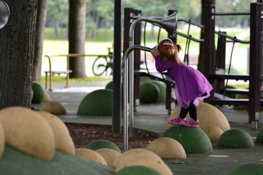 Kırmızı saçlı, mor elbiseli, pembe sandaletli, yeşil çocuk parkında dengede duran, metal oyun tüpüne konuşan, ekipmanla gölgelenmiş park alanına bakan kız..