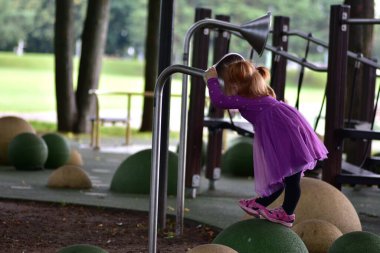 Kırmızı saçlı, mor elbiseli, pembe sandaletli, yeşil çocuk parkında dengede duran, metal oyun tüpüne konuşan, ekipmanla gölgelenmiş park alanına bakan kız..
