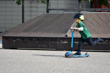 Siyah kask takan genç bir çocuk kaykay parkında mavi üç tekerlekli bir scooter sürüyor, pürüzsüz beton yüzey boyunca ilerlerken konsantre oluyor..