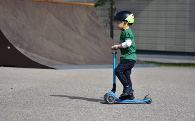 Siyah kask takan genç bir çocuk kaykay parkında mavi üç tekerlekli bir scooter sürüyor, pürüzsüz beton yüzey boyunca ilerlerken konsantre oluyor..