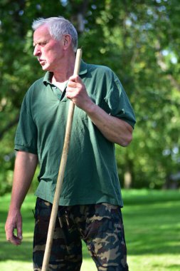 Yeşil gömlekli ve kamuflaj pantolonlu yaşlı bir adamın yakın plan portresi. Elinde bir çubuk, arka planda yeşil ağaçlarla parlak bir kırsal alanda düşünceli görünüyor..