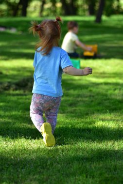 Mavi gömlekli ve çiçekli taytlı kızıl saçlı çocuk arka planda oyuncak kamyonlarla oynayan başka bir çocuğa doğru koşuyor güneşli bir bahçede oyun saatinin keyfini çıkarıyor..