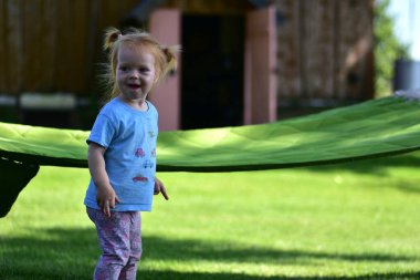 Mavi gömlekli ve desenli taytlı kırmızı saçlı gülen bir kız. Güneşli bir kırsalın arka bahçesinde açık yeşil bir hamağın yanında yeşil çimlerin üzerinde duruyor..