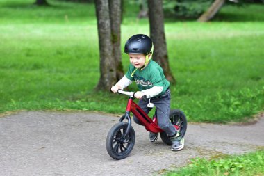 Park yolunda kırmızı dengeli bisiklet süren, güvenlik için kask takan gülümseyen bir çocuk. Aktif çocuk dışarıda eğlenmeyi, yeşil bir çevrede bağımsız olarak dengelemeyi ve dönmeyi öğrenmeyi seviyor..