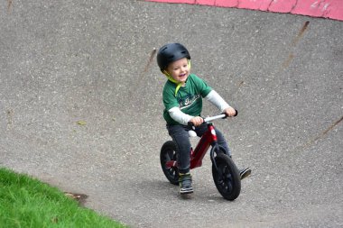 Kavisli asfalt pistte kırmızı dengeli bisiklete binen mutlu çocuk yeşil gömlek ve güvenlik kaskı takıyor parkta açık hava aktivitelerinin, dengenin ve koordinasyon eğitiminin keyfini çıkarıyor..