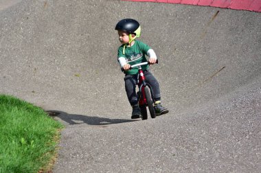 Kırmızı dengeli bisiklete binen çocuk yeşil çimenli eğri bir pistte, kask ve yeşil gömlek giyiyor, dışarıda bisiklet sürerken eğleniyor, spor yapıyor ve çocuklukta beceri eğitimi alıyor..
