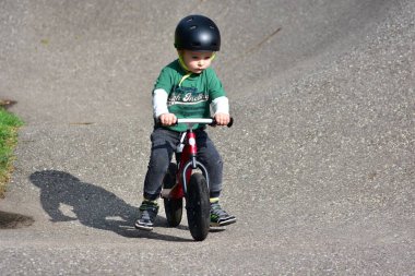 Küçük çocuk asfalt pistinde kırmızı dengeli bisiklete biniyor, miğfer ve yeşil gömlek giyiyor, odaklanmış yüz, denge ve motor becerilerini geliştiriyor, açık havada oyun zamanından ve bisiklet eğitiminden keyif alıyor..