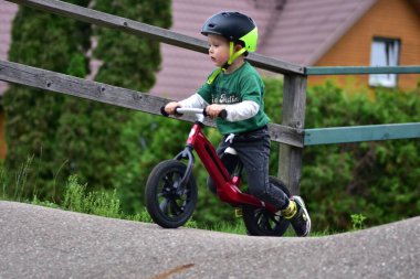 Neon kasklı, yeşil tişörtlü, kırmızı dengeli bisiklete binen küçük çocuk. Odaklanmış çocuk bisiklet öğreniyor ve park ortamında açık havada eğleniyor..