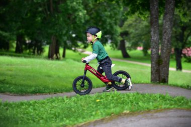 Park yolunda kırmızı dengeli bisiklet süren, güvenlik için kask takan gülümseyen bir çocuk. Aktif çocuk dışarıda eğlenmeyi, yeşil bir çevrede bağımsız olarak dengelemeyi ve dönmeyi öğrenmeyi seviyor..