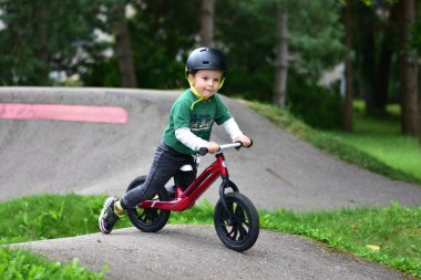 Kask takan küçük çocuk asfalt tulumba pistinde kırmızı dengeli bisiklet sürüyor. Aktif çocuk parkta oynarken denge ve koordinasyon öğreniyor.