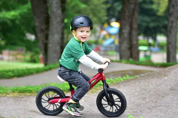 Kask takan küçük çocuk asfalt tulumba pistinde kırmızı dengeli bisiklet sürüyor. Aktif çocuk parkta oynarken denge ve koordinasyon öğreniyor.