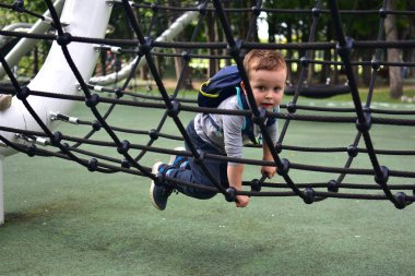 Boy with backpack climbing rope net on playground. Childhood adventure, courage, and physical activity outdoors in park environment.