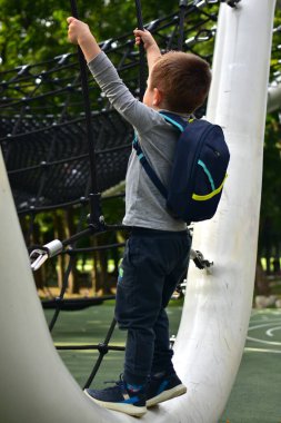 Boy with backpack climbing up ropes on playground structure. Childhood activity, courage, and fun exercise in outdoor park setting.