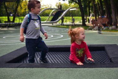 Boy with backpack and girl in red sweater playing together on trampoline in playground. Fun childhood, siblings, and outdoor lifestyle moment.