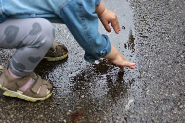 Küçük kızın asfalttaki su birikintisine dokunuşunun yakın çekimi. Çocuk yağmurdan sonra suyu keşfediyor, dışarıdaki merakın ve eğlencenin tadını çıkarıyor.