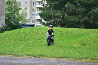 Young boy riding red balance bike downhill on grassy slope. Child wearing helmet, enjoying active outdoor play near apartment buildings. Healthy lifestyle for kids.