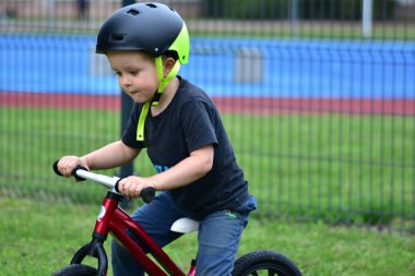 Focused young boy on red balance bike, wearing protective helmet. Close-up portrait of child riding outdoors on green grass. Active childhood sport and learning.
