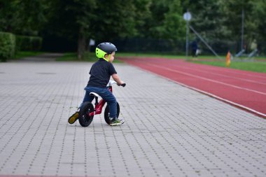 Young boy riding red balance bike on paved track near stadium. Child with helmet enjoying sport activity, practicing balance outdoors. Childhood exercise concept.