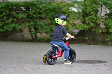 Rear view of boy riding red balance bike on paved square. Child wearing black shirt and green sneakers with helmet. Active outdoor play and healthy childhood lifestyle.