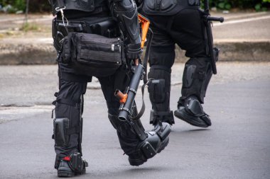 UNDEMO biriminden iki Kolombiyalı isyan karşıtı polis memurunun alt ve ayaklarının bir sokakta yürüdüğünü gösteren bir editör fotoğrafı. İçlerinden biri göz yaşartıcı bomba taşıyor. Görünür yüz yok