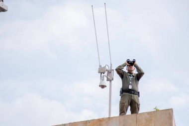 11 Haziran 2025 'te Kolombiya' nın Cali kentini izlerken bir binanın çatısında duran özel bir güvenlik görevlisini gösteren editör fotoğrafı. Arka planda bir güvenlik kamerası görünüyor.