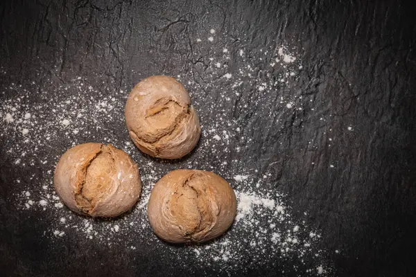 Üzerinde un tozu olan koyu taştan bir zemin üzerinde üç tane sade ekmek rulosu. Esnaf stili mini ekmekler minimum düzeyde, üstüste konulmuş. Fırın, köy yemekleri ve doğal malzemeler için ideal..