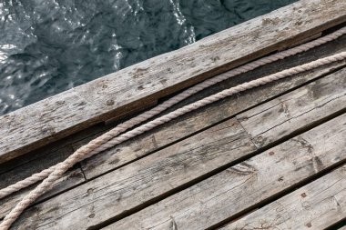 Tahtalara paralel uzanan görünür halatı olan eski ahşap iskelenin bir kısmı, arka planda kısmi su yüzeyi, minimalist ve yıpranmış dış yapı