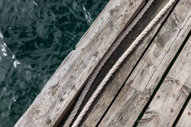 Tahtalara paralel uzanan görünür halatı olan eski ahşap iskelenin bir kısmı, arka planda kısmi su yüzeyi, minimalist ve yıpranmış dış yapı