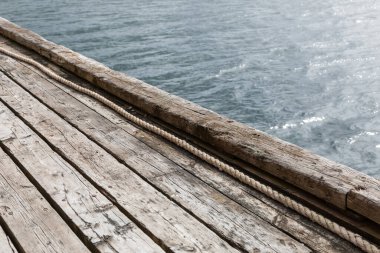 Tahtalara paralel uzanan görünür halatı olan eski ahşap iskelenin bir kısmı, arka planda kısmi su yüzeyi, minimalist ve yıpranmış dış yapı