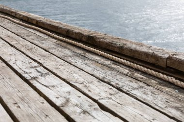 Tahtalara paralel uzanan görünür halatı olan eski ahşap iskelenin bir kısmı, arka planda kısmi su yüzeyi, minimalist ve yıpranmış dış yapı