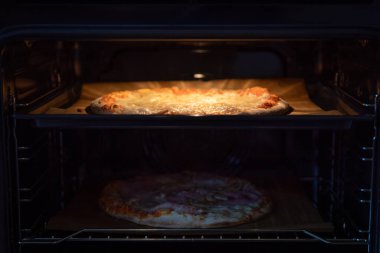 Fırının içinde iki pizza, fırın ışığıyla aydınlatılmış parşömen kağıdıyla pişirme tepsisi, gölgede parşömen kağıdıyla alt rafta, açık fırın kapısından önden vurulmuş.