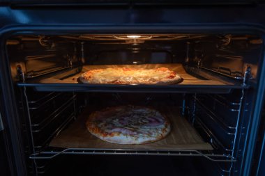 İki donmuş pizzanın ön görüntüsü fırının içinde açık kapılı, biri fırın ışığıyla aydınlatılmış, diğeri gölgede, fırının içi pizzalarla çevrili.
