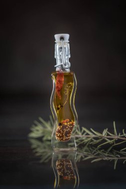 Yansıtıcı yüzeyde baharatlı yağ şişesi ve biber içeren dikey yemek fotoğrafçılığı, yanında taze biberiye, karanlık stüdyo arka planı.