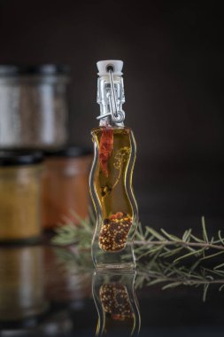 Yansıtıcı yüzeyde baharatlı yağ şişesi ve biber içeren dikey yemek fotoğrafçılığı, yanında taze biberiye, karanlık stüdyo arka planında bulanık üç baharat kavanozu.  
