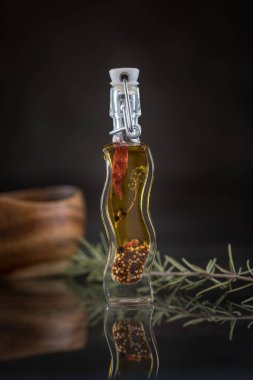 Yansıtıcı yüzeyde baharatlı yağ şişesi ve biber içeren dikey yemek fotoğrafçılığı yanında taze biberiye, karanlık stüdyo arka planında bulanık ahşap bardak.  