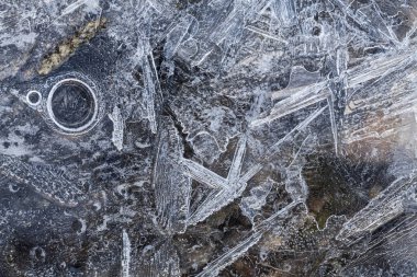 Büyük buz kristalleri ve yüzeyin altındaki donmuş hava kabarcığıyla kıyı buzunun makro yukarıdan aşağı görüntüsü, şeffaf tabakanın altında soluk taşlar, desenli donmuş detaylarla doğal soyut desenler ortaya çıkarıyor.