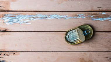 Yıpranmış bej ahşap masa üzerinde yarı açık teneke balık konservesi, kabuk soyulmuş boya, kırsal dokuyu ve metalik yüzeyi gösteren tepeden tırnağa düz bir stüdyo resmi..