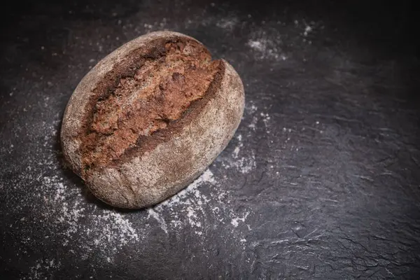 Koyu levha yüzeyinde çatlak kabuklu tam ekşi ekmek, hafif un tozuyla kaplanmış, karamsar ışıklandırma ile kırsal stüdyoda fotoğraflanmış.