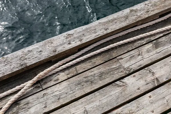 Tahtalara paralel uzanan görünür halatı olan eski ahşap iskelenin bir kısmı, arka planda kısmi su yüzeyi, minimalist ve yıpranmış dış yapı