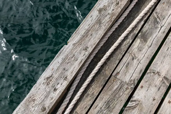 Tahtalara paralel uzanan görünür halatı olan eski ahşap iskelenin bir kısmı, arka planda kısmi su yüzeyi, minimalist ve yıpranmış dış yapı
