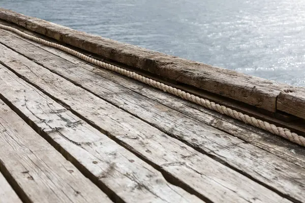 Tahtalara paralel uzanan görünür halatı olan eski ahşap iskelenin bir kısmı, arka planda kısmi su yüzeyi, minimalist ve yıpranmış dış yapı
