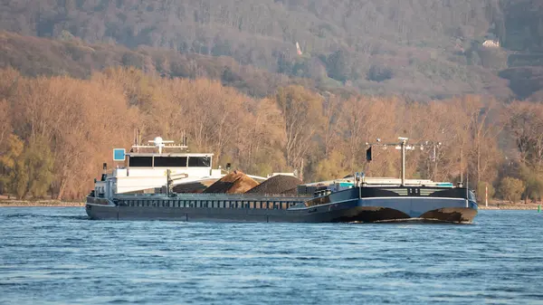 Sonbaharda Ren nehrinin nehir seviyesine bakın. Malzeme taşıyan kargo gemisi, önplanda bulanık mavi su ve akşam güneşinin altında sonbahar renkli ormanlar ve tepeler.