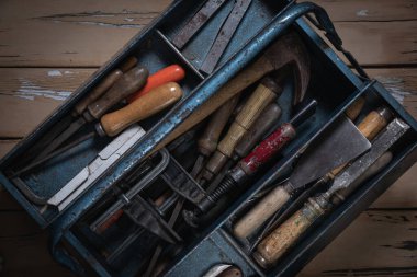 Eski bej boyalı ahşap masa ve yontulmuş boya, merkez olarak yerleştirilmiş mavi metal alet kutusu ana bölme ile çapraz konum ve üst iki tepsi tamamen açık, içerideki çeşitli eski araçları gösteren.