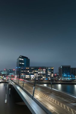 Gece boyunca Hamburg HafenCity 'deki Baakenhafen Köprüsü' nde aydınlık Versmannkai rıhtımı ve sakin suya yansıyan koyu mavi gökyüzü..  