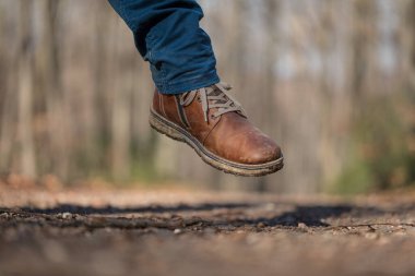 Yakın plan kahverengi deri yürüyüş botu sonbahar orman patikasının üzerinde, bacağı mavi pantolonlu bacağa kadar görünür, ayakkabı yüzeyindeki kir, bulanık ağaçlar ve arka plandaki orman yolu.  