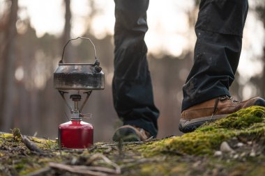 Person standing beside camping stove with kettle on forest floor, low angle view showing legs and hiking boots, outdoor daytime scene  