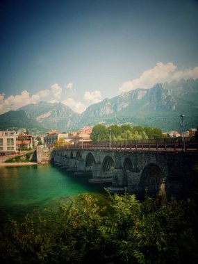 Nehir boyunca uzanan tarihi bir taş köprü, yeşilliklerle çevrili.