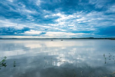 Kabini 'nin durgun sularının sakin manzarası dramatik bulutları yansıtan cam gibi bir yüzeyi var. Nadir bitki örtüsü ve uzak tekneler huzurlu ve doğal atmosferi güçlendirir..